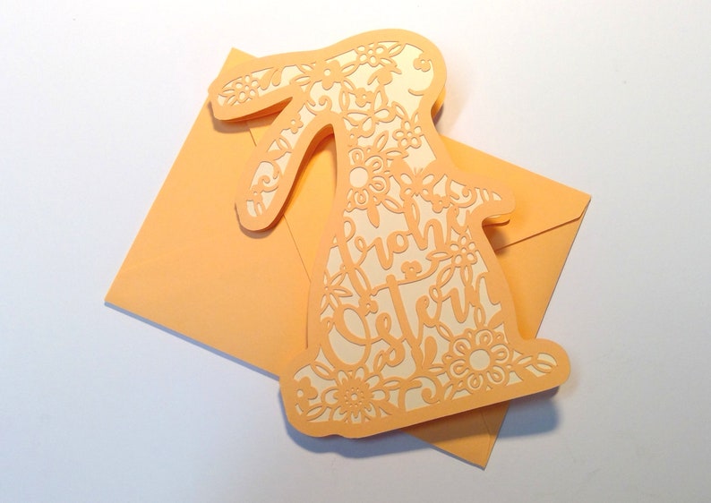 May include: A peach-colored card with a white bunny cutout. The bunny is decorated with floral designs and the text "Frohe Ostern" is inside the cutout.
