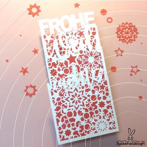 May include: A rectangular white paper cutout card featuring intricate laser-cut patterns of snowflakes, stars, and circles. The text FROHE WEIHNACHTEN is cut out in a bold, stylized font. The card is layered over a solid red background.