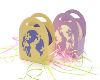 May include: Two Easter gift boxes with handles, one yellow and one purple, with a cut-out design of a bunny rabbit in a field of flowers. The boxes are filled with pink and green shredded paper.