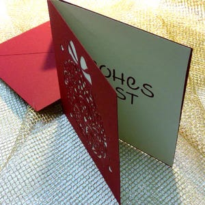 May include: A red greeting card with a laser-cut floral pattern on the front cover and a matching red envelope. The interior of the card features the visible text "WISHES BEST" printed in a dark font against a light cream-colored background.