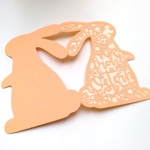 May include: A peach-colored card with two cutout bunny silhouettes. The bunny on the right has a floral design and the text "Happy Easter" inside the cutout.