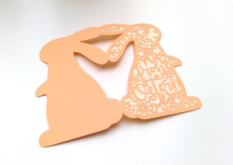 May include: A peach-colored card with two cutout bunny silhouettes. The bunny on the right has a floral design and the text "Happy Easter" inside the cutout.
