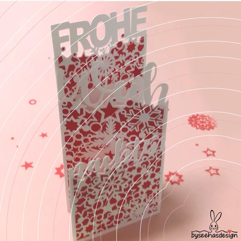 May include: A vertical greeting card featuring a white laser-cut overlay with intricate snowflake and star patterns over a solid red background. The top of the card displays the text FROHE in white block letters, with additional script text below.