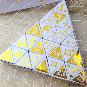 May include: A triangular advent calendar composed of individual white and gold triangular boxes. Each box features a number from 1 to 25 in gold foil, with some boxes displaying German number words like ZWEI, ACHT, NEUN, SECHZEHN, and ZWANZIG.