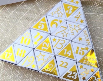 May include: A triangular advent calendar composed of individual white and gold triangular boxes. Each box features a number from 1 to 25 in gold foil, with some boxes displaying German number words like ZWEI, ACHT, NEUN, SECHZEHN, and ZWANZIG.