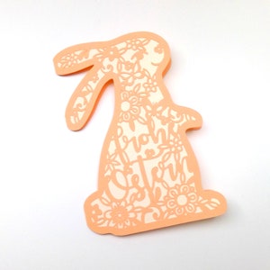 May include: A peach-colored paper cutout of a bunny rabbit with intricate floral designs and the text "Frohe Ostern" inside the bunny's body.