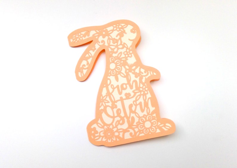 May include: A peach-colored paper cutout of a bunny rabbit with intricate floral designs and the text "Frohe Ostern" inside the bunny's body.