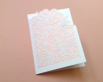 May include: A white card with a delicate floral design cut out, revealing a light pink background.
