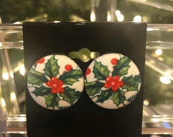 May include: A pair of round stud earrings with a white background, green leaves, and red berries pattern. The earrings are displayed on a black card, perfect for holiday or Christmas gifts. The earrings are a festive accessory.