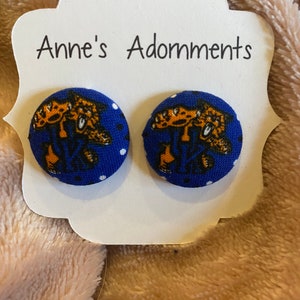 May include: A pair of button earrings with a blue background and a white polka dot pattern. Each earring features a gold and black graphic design. The earrings are displayed on a white card with the text "Anne's Adornments".
