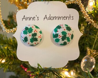 May include: A pair of white button earrings with a green and red holly pattern. The earrings are displayed on a white card with the text "Anne's Adornments".