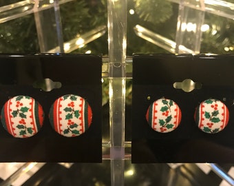 May include: Button earrings with a festive Christmas design. The earrings feature a white background with a holly and berry pattern, framed by red and green stripes. The earrings are displayed on black earring cards.