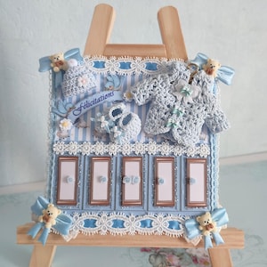 May include: A light blue decorative easel with baby-themed embellishments. Features a crocheted sweater, hat, and booties, along with a banner that reads "Félicitations". Includes decorative bows and teddy bears.