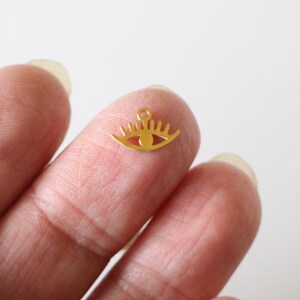 Pode incluir: Um pequeno pingente dourado em forma de olho. O pingente tem uma argola na parte superior para prender a uma corrente ou outro componente de joia. O olho tem um design estilizado com cílios.