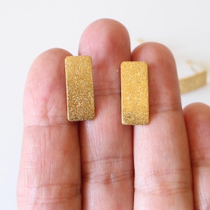 Peut inclure: Une paire de boucles d'oreilles clous rectangulaires dorées avec une surface texturée et scintillante. Les boucles d'oreilles sont présentées sur une main sur fond blanc. Design simple et élégant.