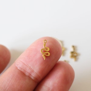 Pode incluir: Pingente de cobra em tom dourado com um laço na parte superior para prender em joias. O charme mede aproximadamente 2,5 cm de comprimento e é mostrado em um dedo.