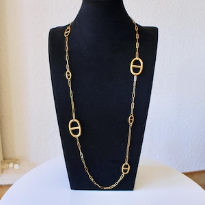 Puede incluir: Un collar de cadena larga en tono dorado que presenta una serie de eslabones ovalados alargados intercalados con dijes ovalados horizontales más grandes que contienen una barra central, exhibido en un busto de terciopelo negro.