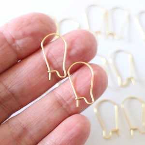 May include: Gold-toned earring hooks, a close-up of two hooks held in a hand, with several more hooks scattered on a white surface.