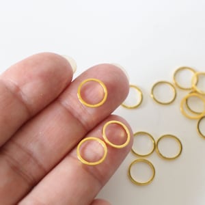 Peut inclure: Gros plan sur une main tenant plusieurs petits anneaux de jonction dorés. Les anneaux mesurent environ 5 mm de diamètre et sont parfaits pour la fabrication de bijoux.
