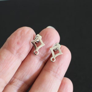 Pode incluir: Dois achados de brincos prateados com um design em forma de diamante. Cada achado tem um laço na parte inferior para prender um amuleto ou um pingente.