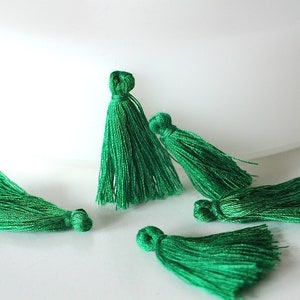 Puede incluir: Cinco borlas verde esmeralda de diferentes tamaños dispuestas sobre un fondo blanco. Cada borla está hecha de hilos brillantes y apretados, con una parte superior en bucle y una parte inferior con flecos. Las borlas son probablemente para manualidades o decoración.