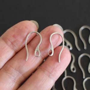 Op de afbeelding: Twee zilveren oorhaken, met een kleine lus aan de bovenkant, worden in een hand gehouden. De haken zijn van metaal en zijn bedoeld om oorbellen aan te bevestigen.