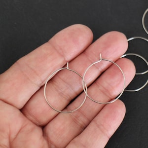 May include: A close-up of a hand holding two silver hoop earrings. The earrings are simple and circular in shape. There are several other silver hoop earrings in the background.