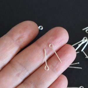 Puede incluir: Alfileres de cabeza plateada con un bucle en la parte superior. Los alfileres miden aproximadamente 2,5 cm de largo y están esparcidos sobre una superficie negra.