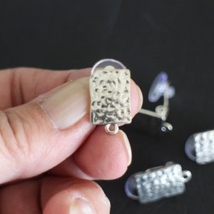 Peut inclure: Fermoirs de boucles d'oreilles argentés avec une texture martelée et une boucle pour attacher un pendentif. Les fermoirs sont présentés sur une surface noire.