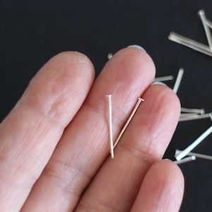 Peut inclure: Gros plan sur une main tenant deux épingles à tête argentées. Les épingles mesurent environ 2,5 cm de long et ont une tête arrondie. Il y a beaucoup d'autres épingles à tête argentées éparpillées sur une surface noire.