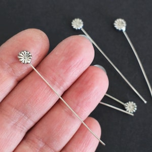 Op de afbeelding: Zilveren bloemkopspelden, met een decoratief bloemmotief op de kop van elke speld. De spelden zijn dun en recht en worden in een hand getoond.