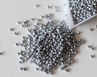 May include: A large pile of silver seed beads, scattered on a white surface. The beads are small and round, with a metallic sheen.