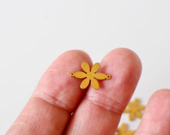 May include: A small, gold-colored metal flower charm with six petals and two small holes for attaching to jewelry. The charm is held between fingers against a white background.