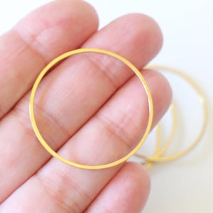 Pode incluir: Vários brincos de argola de metal dourado são exibidos. Os aros são finos e circulares, com um acabamento liso e brilhante. Os brincos são segurados em uma mão contra um fundo branco. Adequados para fazer joias.