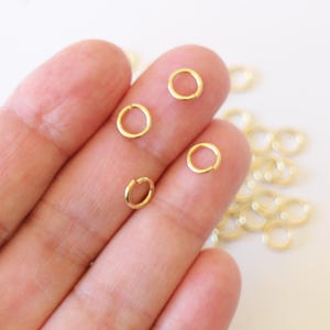 Op de afbeelding: Een close-up van een hand die vier gouden springringen vasthoudt. De ringen zijn klein en rond en perfect voor het maken van sieraden.