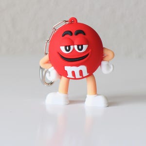 Puede incluir: Llavero con la figura de un M&M rojo con una cara sonriente, ojos blancos y una 'M' blanca en el pecho. El llavero tiene una cadena plateada y zapatos blancos.