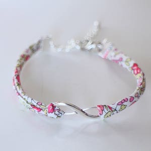 Puede incluir: Un brazalete de tela blanca con estampado floral rosa y verde y un dije de plata en forma de infinito.