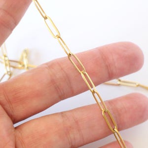 Pode incluir: Uma corrente dourada com elos ovais alongados é segurada em uma mão contra um fundo branco. A corrente é feita de um material metálico brilhante. Os elos são uniformes em tamanho e forma. Joias.