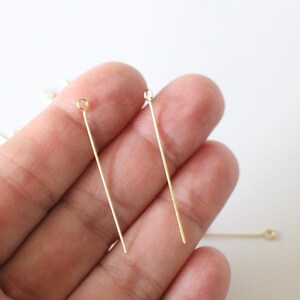 May include: Two gold-coloured head pins with a loop at the top. The pins are held in a hand.