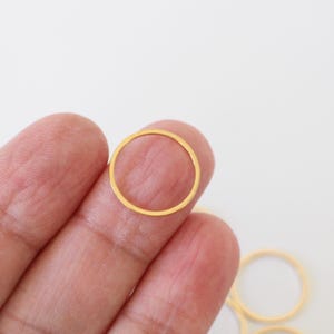 Peut inclure: Gros plan sur un anneau métallique fin et circulaire de couleur or, tenu entre les doigts sur fond blanc. L'anneau est de conception simple et minimaliste, et plusieurs autres anneaux sont visibles.