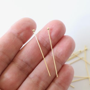 Peut inclure: Deux épingles à tête dorées avec des têtes rondes, tenues dans une main. Un tas d'épingles à tête supplémentaires est visible en arrière-plan.