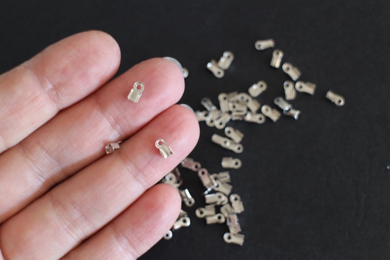 May include: A close-up of a hand holding several silver crimp end caps. The caps are small and have a rounded shape. They are used to secure the ends of cords or strings.