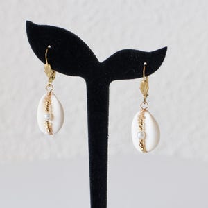 May include: A pair of drop earrings featuring white cowrie shells accented with a vertical line of small gold-toned beads and a single central white pearl. The shells are suspended from gold-toned metal lever-back hooks for secure attachment.