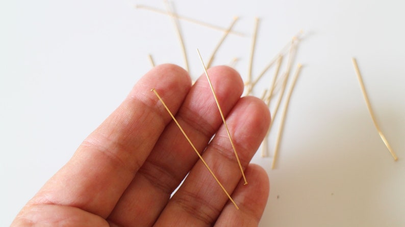 Pu&ograve; includere: Primo piano di una mano che tiene due spilli dorati con la testa. Gli spilli sono sottili e dritti, e misurano circa 2,5 cm di lunghezza. Ci sono altri spilli sparsi sulla superficie bianca.