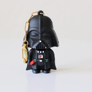 Puede incluir: Un llavero negro de Darth Vader con una cadena y un cierre dorados. La figura tiene un casco y un uniforme detallados, con un sable de luz rojo. El llavero es un artículo de colección de la franquicia Star Wars.