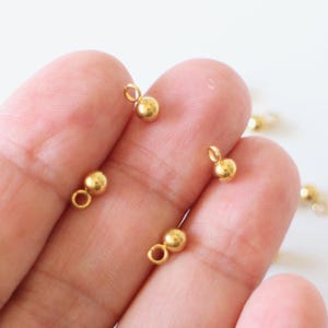 Könnte beinhalten: Nahaufnahme mehrerer kleiner, goldfarbener Schmuckzubehörteile. Jedes Zubehörteil hat eine kleine Schlaufe und eine abgerundete Kugelform. Die Zubehörteile sind auf hellem Hintergrund verstreut.