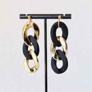 May include: A pair of drop earrings featuring three interlocking oval links. The left earring has a gold, black, and gold link sequence, while the right earring has a black, gold, and black link sequence. Both earrings have gold stud posts.