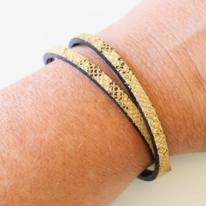 Könnte beinhalten: Ein goldfarbenes und schwarzes Lederarmband. Das Armband besteht aus zwei dünnen Lederstreifen, einem goldenen und einem schwarzen, die um das Handgelenk gewickelt werden.