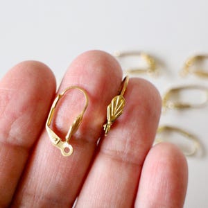 Könnte beinhalten: Nahaufnahme von goldfarbenen Ohrring-Verschlüssen mit Hebelverschluss. Einer ist geöffnet und zeigt Hebel und Schlaufe, während der andere ein dekoratives Blattdesign hat. Im Hintergrund sind weitere Verschlüsse zu sehen.