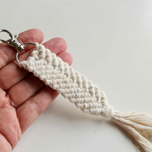 以下が含まれることがあります： 金色の糸が織り込まれた白いマクラメのキーホルダー。シルバーのキーリングと白いタッセルが付いています。